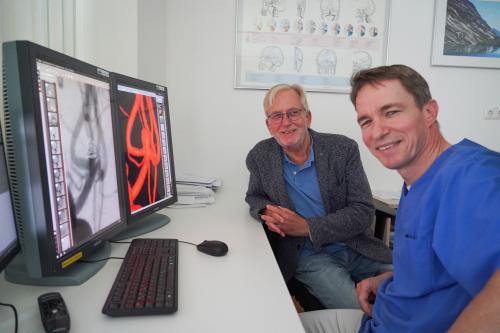 Chefarzt Dr. Christian Loehr (l.) zeigt seinem Patienten Dr. Claus Bardelle (l.) eine vergrößerte Aufnahme des behandelten Hirnaneurysmas. Dr. Loehr und Dr. Bardelle